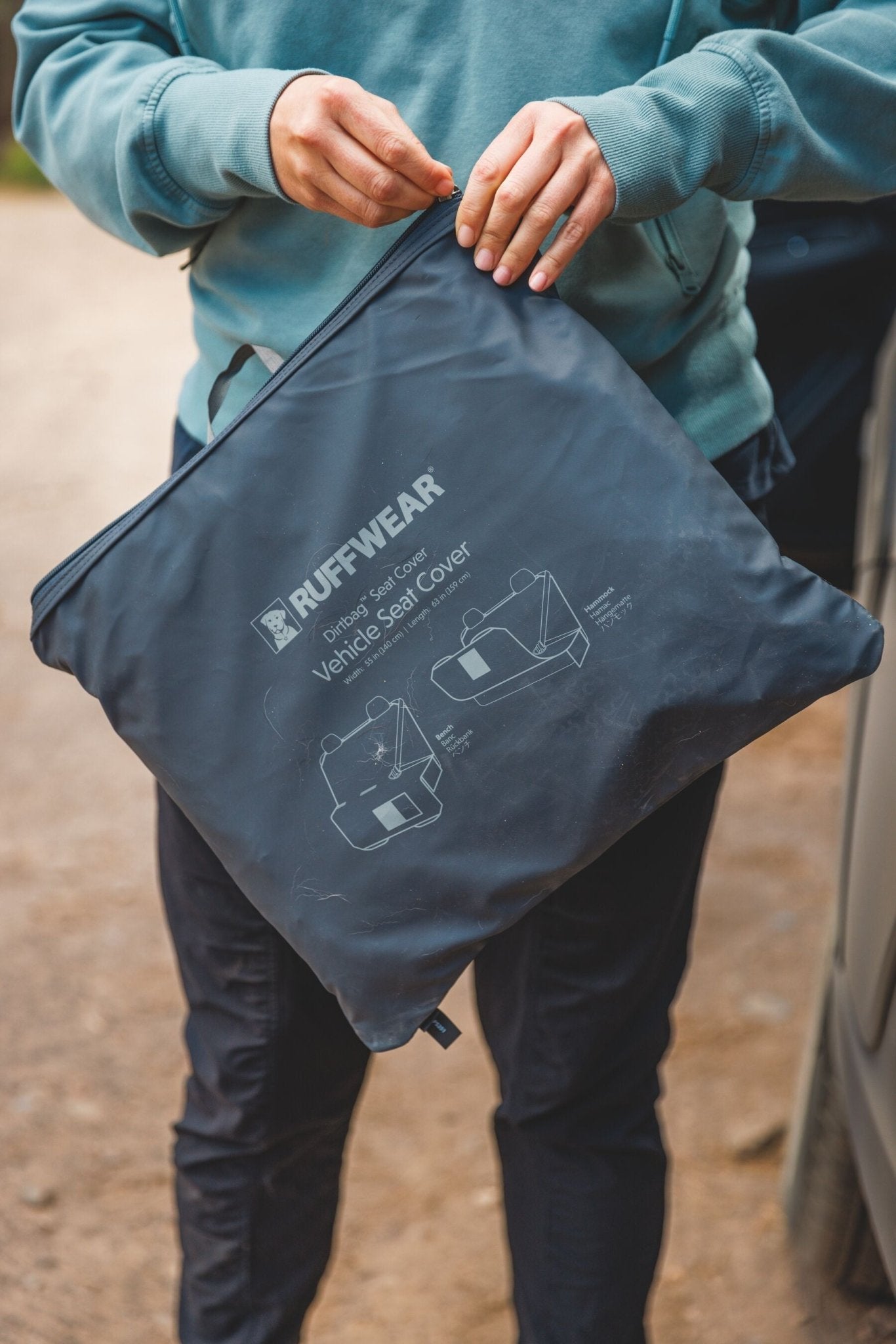 Person in green sweatshirt holding a gray Ruffwear vehicle seat cover bag with a zipper.
