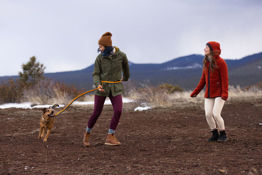 Two women and a dog in an outdoor setting. One woman with a brown beanie and green jacket leads a dog. The other woman wears a red fleece jacket and white leggings.